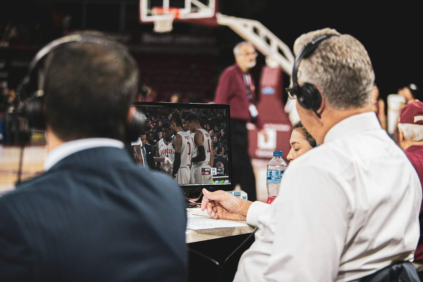 Three sports broadcasters watch a live basketball game on monitors.