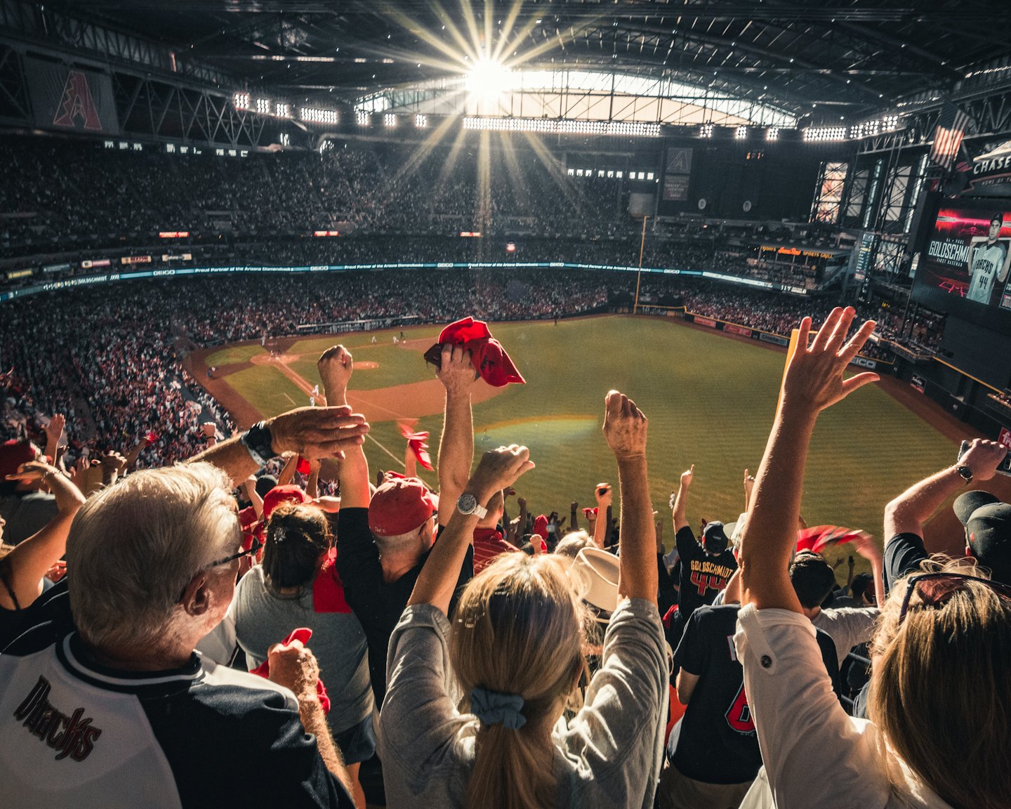 Crowd cheering with arms raised in a sunny baseball stadium.